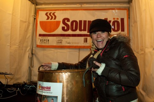 A woman in a winter jacket smiles while holding a large pot at a Soupköl event, with a banner in the background.
