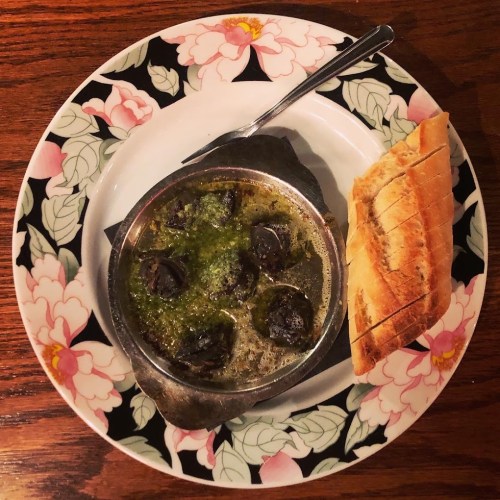 A plate of escargot in garlic butter, served with a slice of bread on the side, set on a floral-patterned plate.