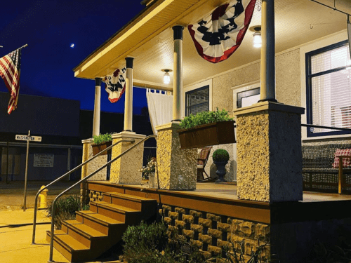A cozy porch at night, decorated with American flags and bunting, featuring steps and potted plants.
