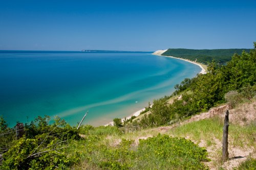 A scenic view of a tranquil blue lake bordered by green hills and sandy shores under a clear blue sky.
