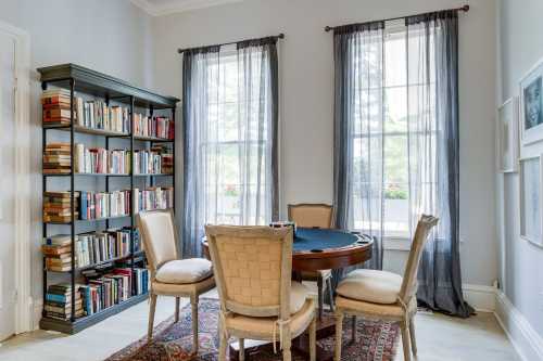 Cozy room with a round table, four chairs, a bookshelf filled with books, and large windows with sheer curtains.