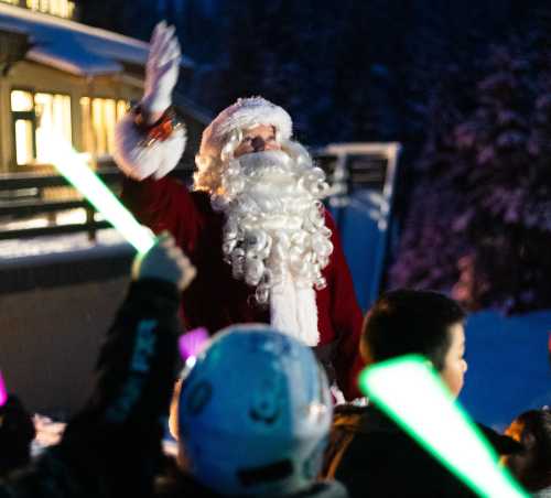 Santa Claus waves to a crowd of children holding colorful glow sticks in a snowy outdoor setting.