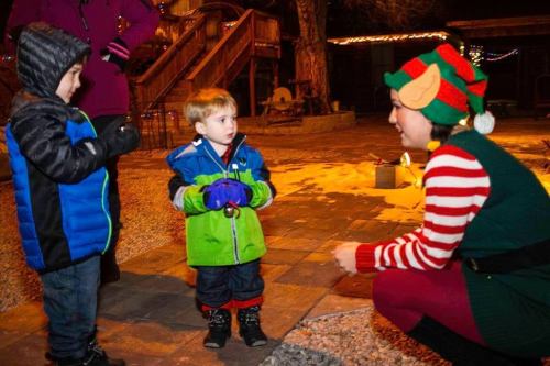 Two young boys in winter jackets interact with a smiling elf in festive attire during a holiday event.
