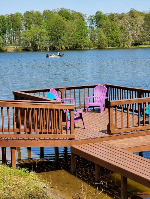 A wooden dock with colorful chairs overlooks a calm lake, where a small boat is seen in the distance.