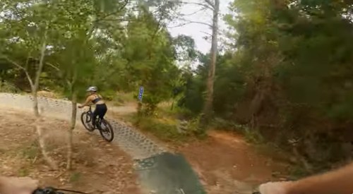 A person rides a mountain bike on a trail surrounded by trees and greenery.