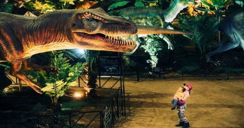 A child and an adult stand in awe before a large dinosaur exhibit in a vibrant, prehistoric-themed environment.