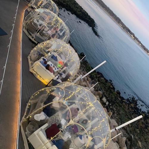 Aerial view of cozy dining pods with lights, overlooking a calm waterway at sunset.