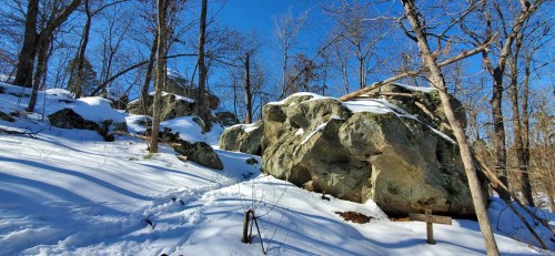 Snow-covered rocky terrain with trees and a clear blue sky in the background. Footprints lead through the snow.