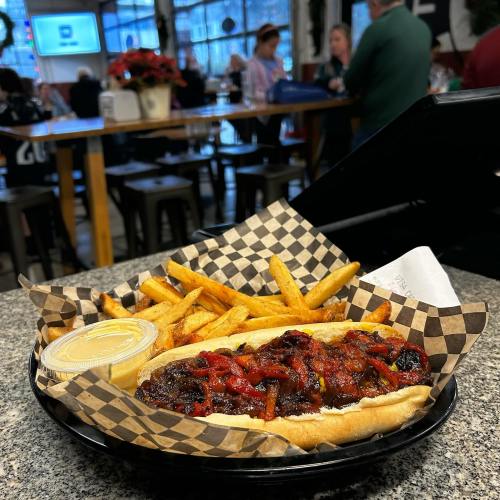 A hot dog topped with chili and peppers, served with fries and dipping sauce, on a checkered paper in a casual dining setting.