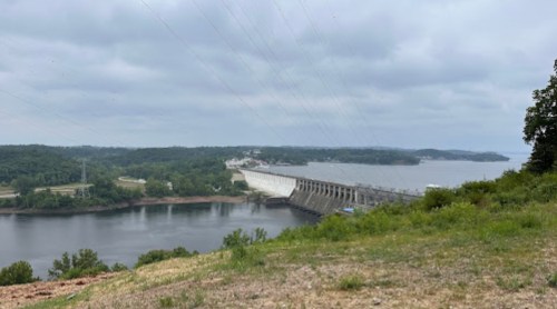 A view of a dam spanning a river, surrounded by green hills and overcast skies.