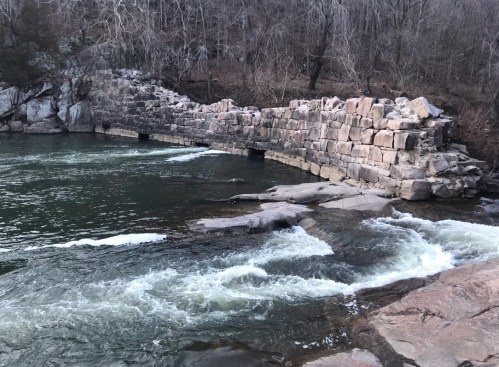 A stone ruin partially submerged in a river, surrounded by trees and rocky banks, with flowing water in the foreground.