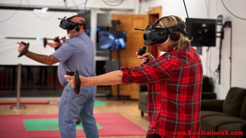 Two people wearing VR headsets are engaged in a virtual reality experience, holding controllers in a gaming space.