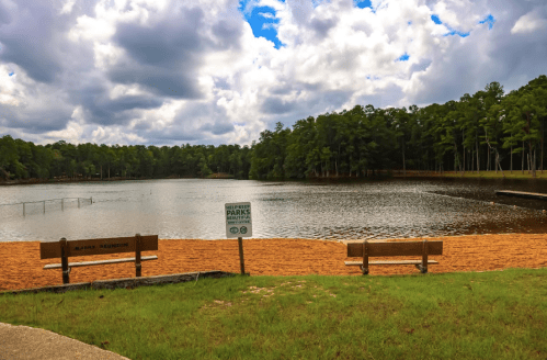 A serene lake surrounded by trees, with sandy beach areas and benches overlooking the water under a cloudy sky.