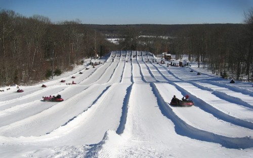A snowy hill with multiple tubing lanes, people sliding down on inflatable tubes under a clear blue sky.