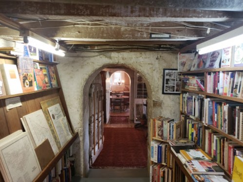 A cozy, dimly lit bookstore hallway with bookshelves on either side and an arched doorway leading to a reading area.