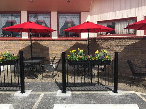Outdoor dining area with black tables and chairs, red umbrellas, and flower planters against a stone wall.