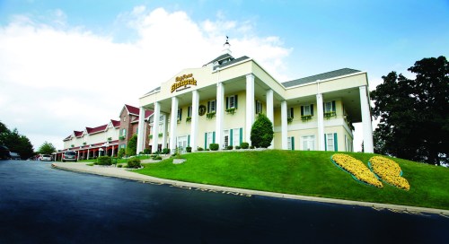 A large, elegant hotel with a manicured lawn and flower arrangements, set against a clear blue sky.