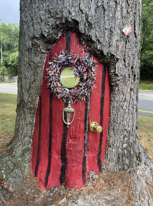 A whimsical red door with a wreath, set into the trunk of a tree, surrounded by a grassy area.