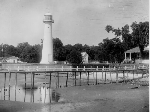 Black and white image of a lighthouse beside a wooden pier, with trees and houses in the background.