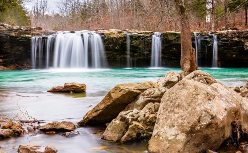 A serene waterfall cascades over rocks into a turquoise pool, surrounded by trees and a tranquil landscape.