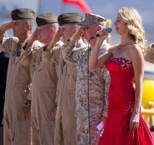 A woman in a red dress sings into a microphone while military personnel salute during a ceremony.