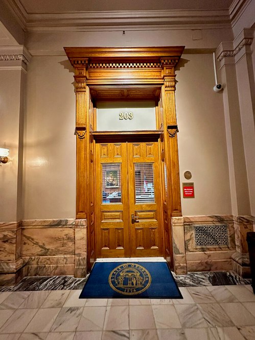 Wooden door with the number 203, framed by ornate architecture, in a marble-floored hallway. Blue mat with a seal in front.