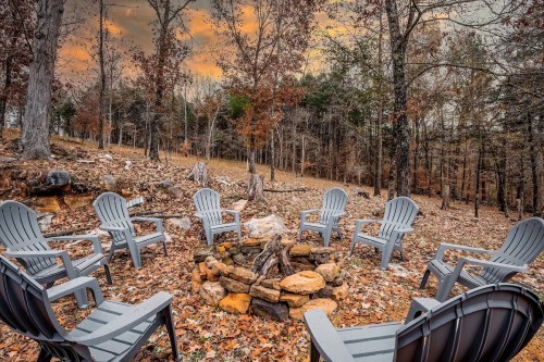 A cozy fire pit surrounded by gray chairs in a wooded area with autumn leaves and a dramatic sky.