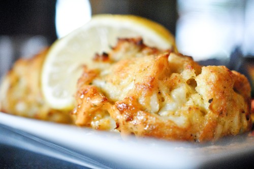 Close-up of a golden-brown crab cake garnished with a lemon slice on a white plate.