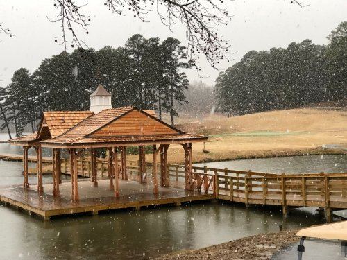 A wooden gazebo by a snowy lake, surrounded by trees and a grassy field, creating a serene winter scene.