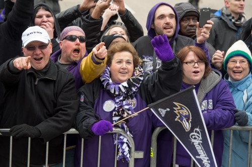 A group of excited fans in purple attire cheer and celebrate, holding a Ravens flag at a public event.