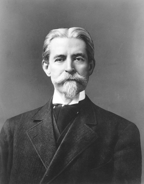 Black and white portrait of a man with a mustache and beard, wearing a formal suit and tie.