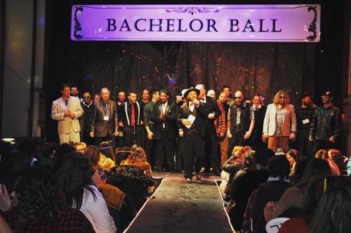 A group of men in formal attire on stage at a Bachelor Ball event, with an audience seated in front.