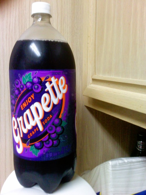 A bottle of Grapette grape soda with a purple label, sitting on a white surface against a wooden background.