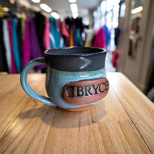 A handcrafted mug with "BRYCE" engraved, resting on a wooden surface, with colorful clothing in the background.