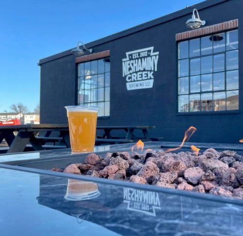 A glass of beer sits on a table with a fire pit, in front of the Neshaminy Creek Brewing Co. building.
