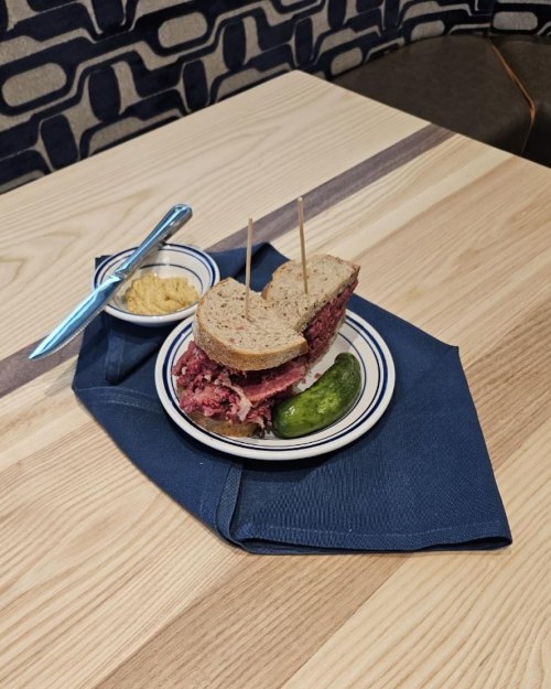 A sandwich with pastrami on rye, served with a pickle and mustard on a blue napkin, on a wooden table.