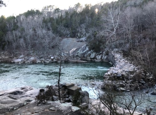 A serene river flows through a rocky landscape, surrounded by bare trees and a distant forested area.