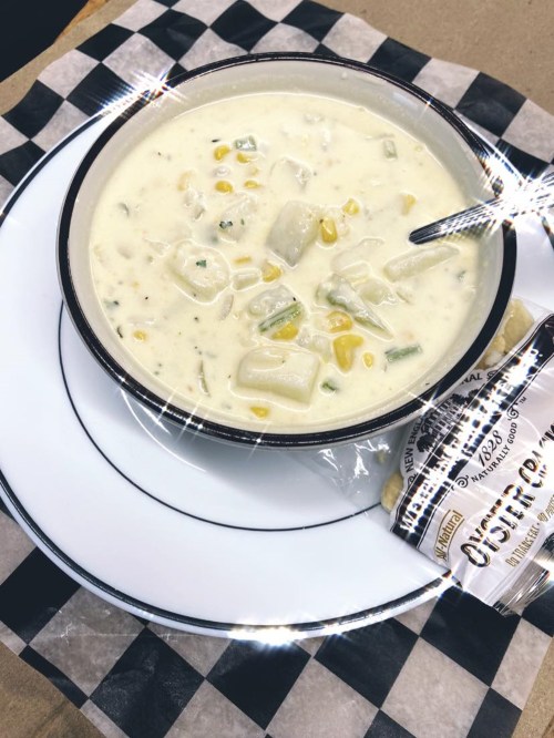 A bowl of creamy corn chowder with potatoes and green onions, served with a packet of oyster crackers on a checkered napkin.