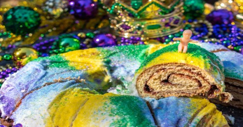 A colorful king cake with green, yellow, and purple icing, topped with a small figurine, surrounded by festive beads.