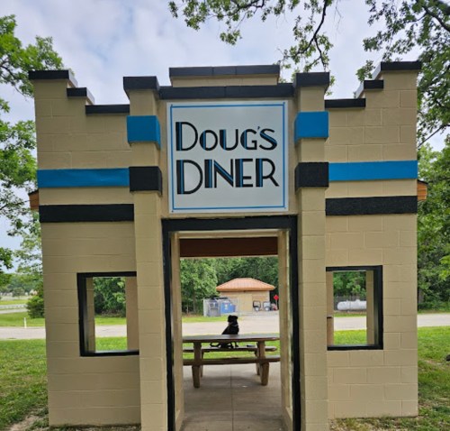 A colorful diner facade with "Doug's Diner" sign, featuring a picnic table in front and trees in the background.