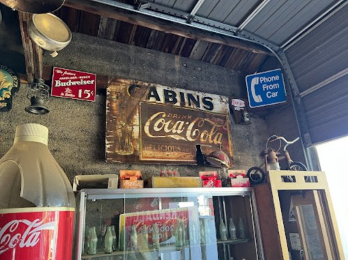 Vintage Coca-Cola sign and memorabilia displayed in a rustic setting, with Budweiser and phone signs in the background.