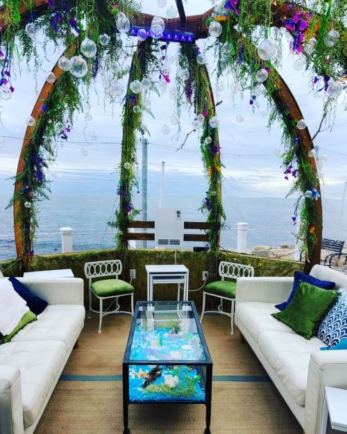 A cozy seating area with white sofas and a glass table, surrounded by greenery and overlooking the ocean.