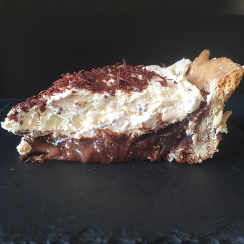 A slice of chocolate cream pie topped with whipped cream and chocolate shavings on a dark slate surface.