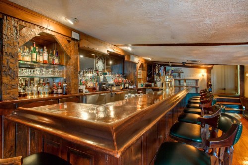 A wooden bar with high stools, shelves of liquor, and a warm, inviting atmosphere in a cozy pub setting.