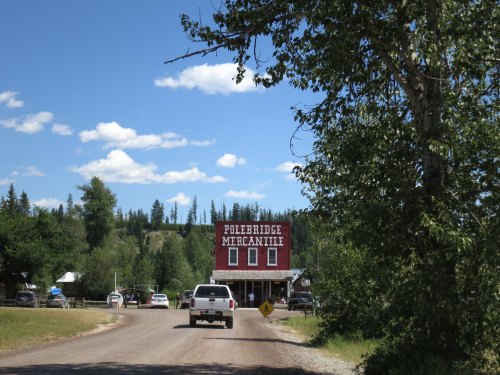 A red mercantile building stands along a dirt road, surrounded by trees and blue skies with scattered clouds.