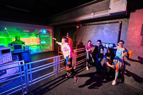 A group of people engaged in an interactive game, aiming toy blasters at a colorful, high-tech screen.