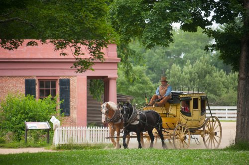 A horse-drawn carriage with a driver passes by a historic building surrounded by greenery.