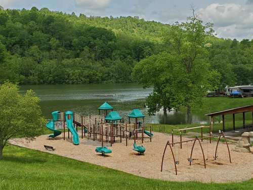 A playground with slides and swings near a lake, surrounded by green trees and a cloudy sky.