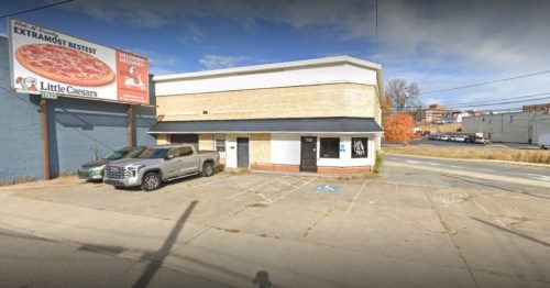 A small commercial building with a black roof, parking lot, and a Little Caesars billboard in the background.