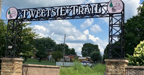Sign for Tweetsie Trail with a blue sky and clouds in the background, surrounded by greenery and buildings.
